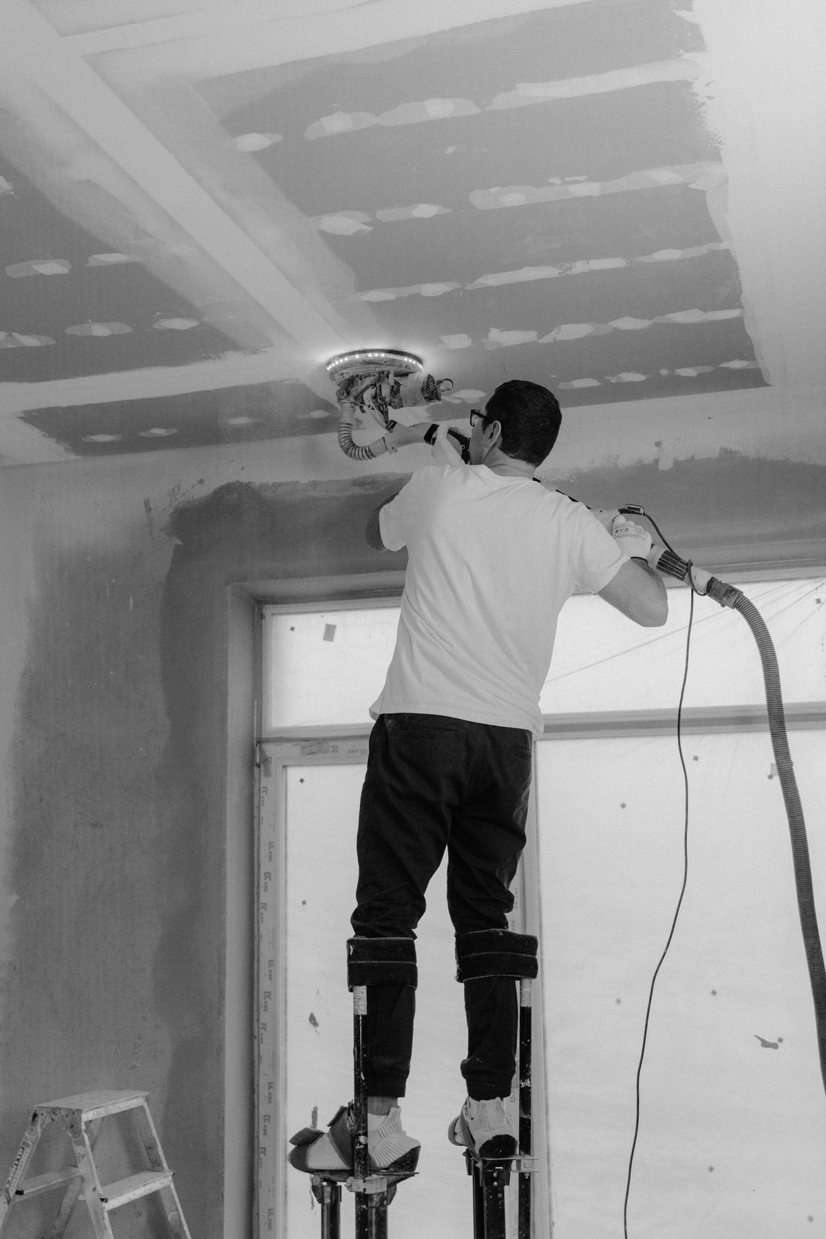 Tradesman on drywall stilts running a sander with an LED ring across a bare ceiling