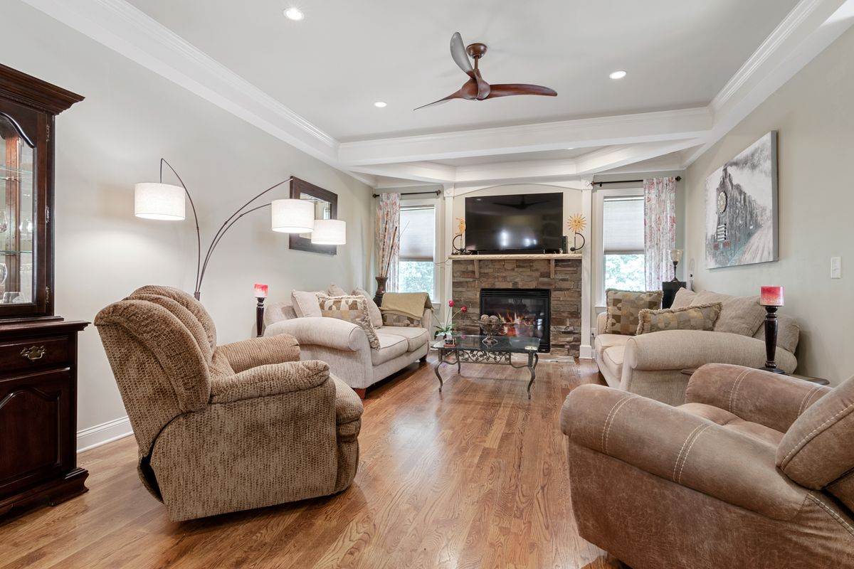 Finished living room with a smooth tray ceiling, crown molding, ceiling fan, and stone fireplace