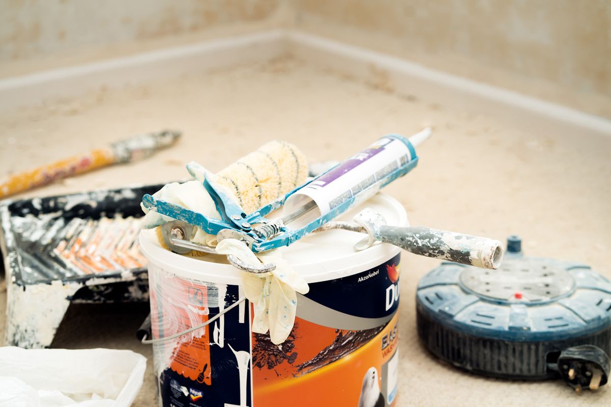 Paint bucket with a roller sleeve, caulk gun, and used brushes resting on it during a renovation
